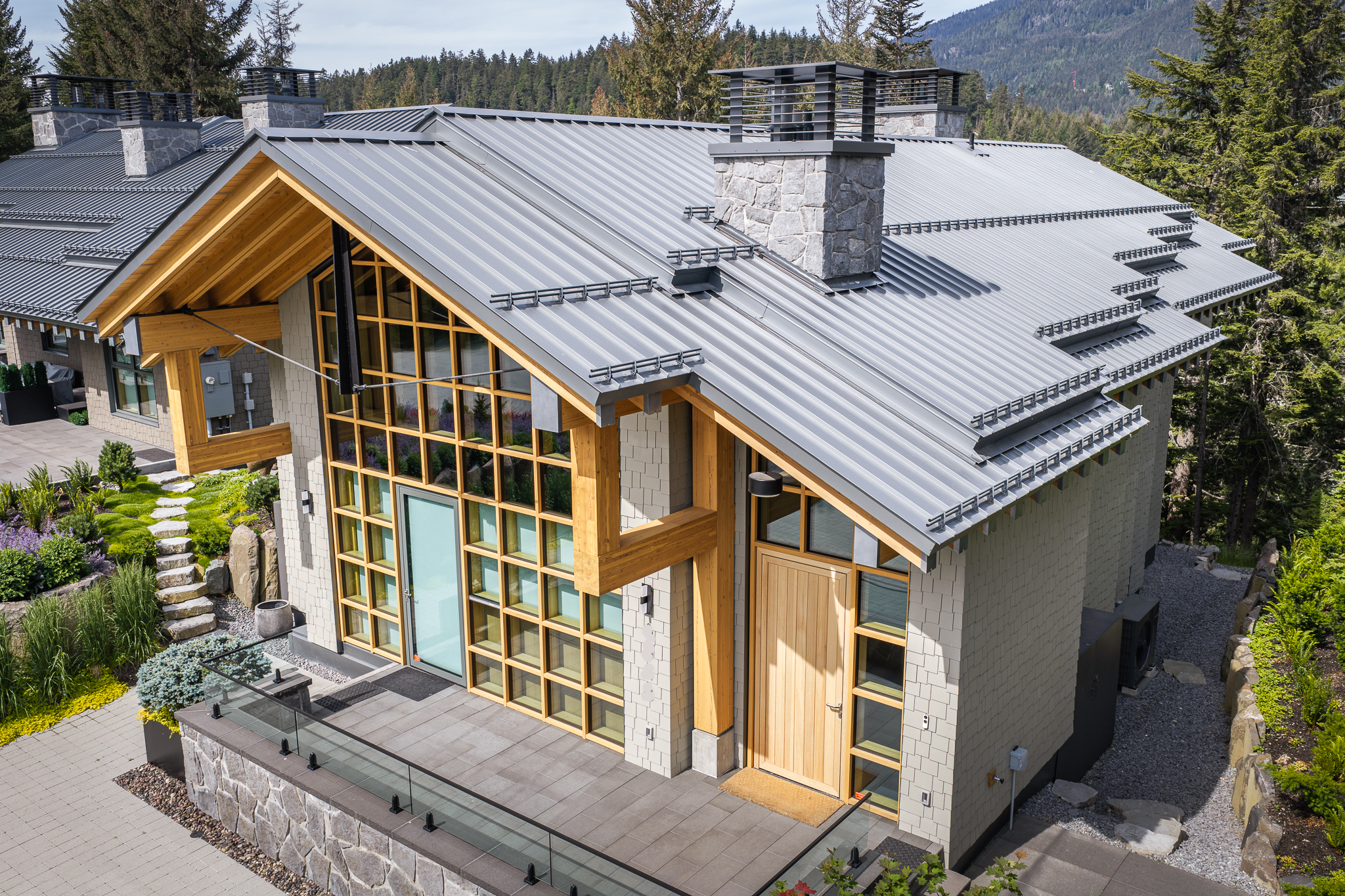 Modern mountain home featuring a standing seam metal roof, large windows, timber accents, and natural stone details, surrounded by landscaped gardens and mature trees.