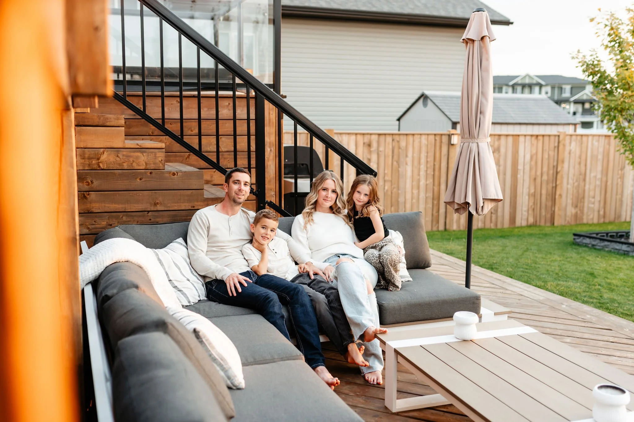 A family of four sitting on a gray outdoor sofa on a wooden deck, with a wooden staircase and railing behind them, and a beige patio umbrella to the right. They are in a backyard with a wooden fence and neighboring houses visible in the background.