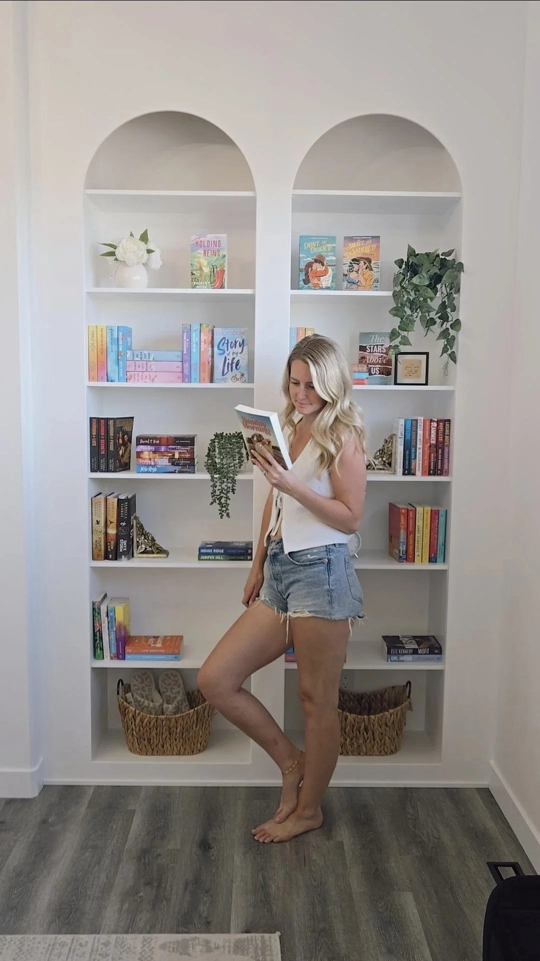A woman with long blonde hair wearing a white sleeveless top and denim shorts standing barefoot in front of a white bookshelf filled with colorful books and decorative items, holding a book and reading.