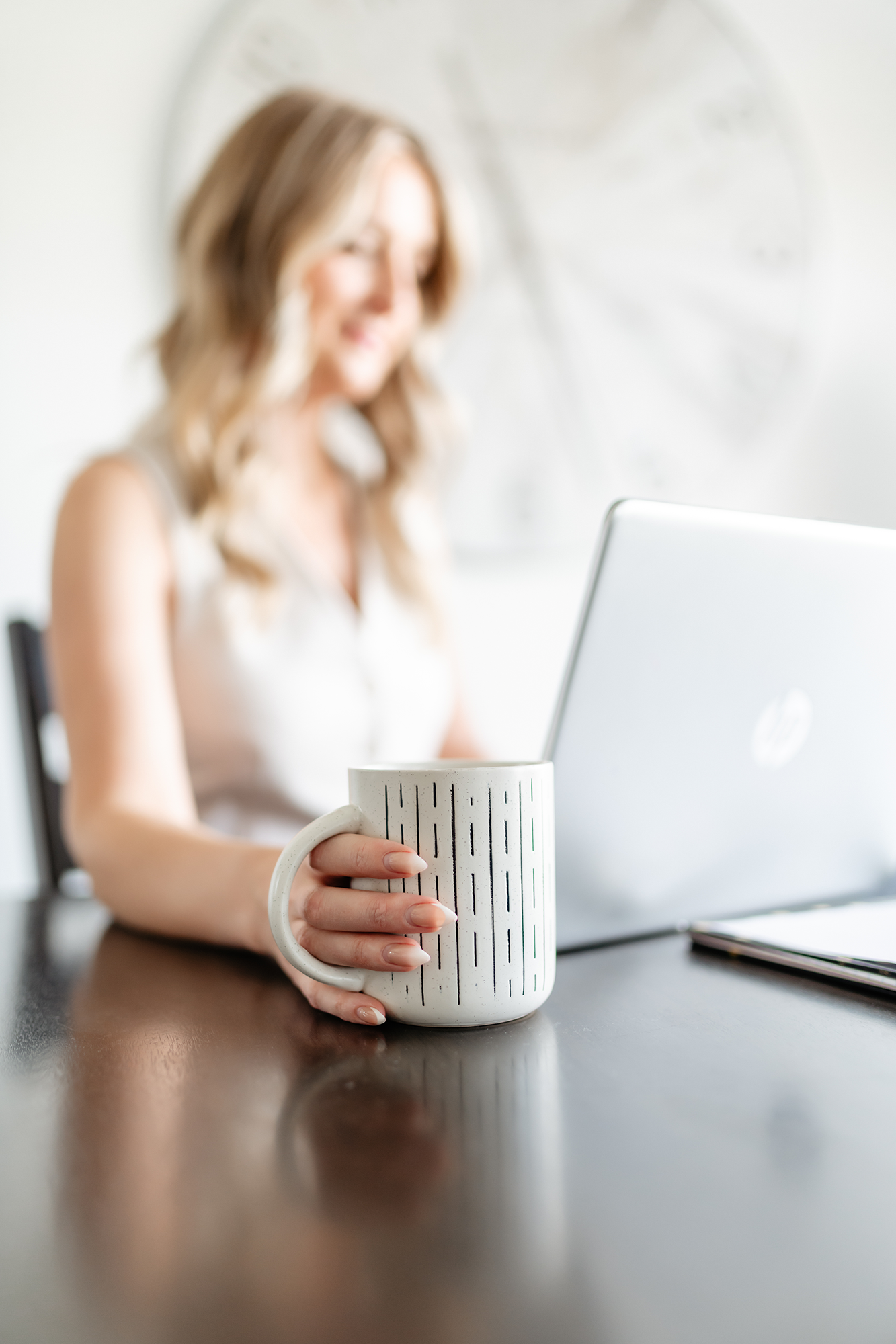 A woman sitting at a table holding a white mug with black and green vertical stripes, with a laptop and a smartphone on the table in front of her, in a bright room with a clock on the wall.