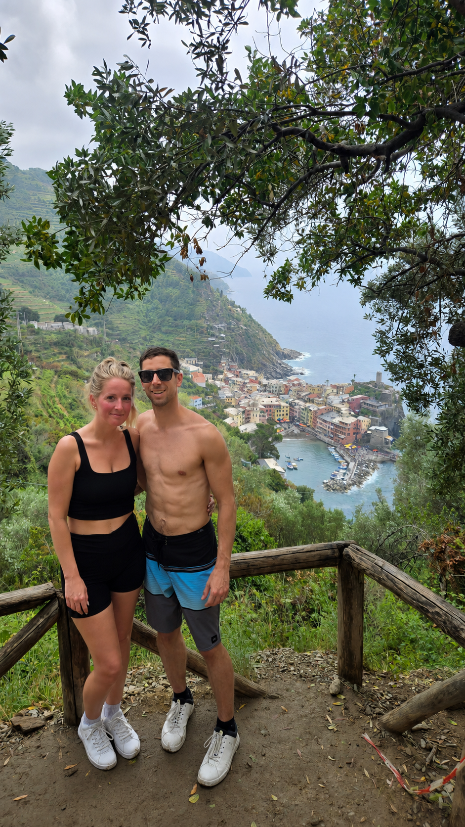 A couple stands on a dirt and wood railing overlooking a coastal village with colorful buildings, a harbor, and the sea, framed by tree branches and green foliage.