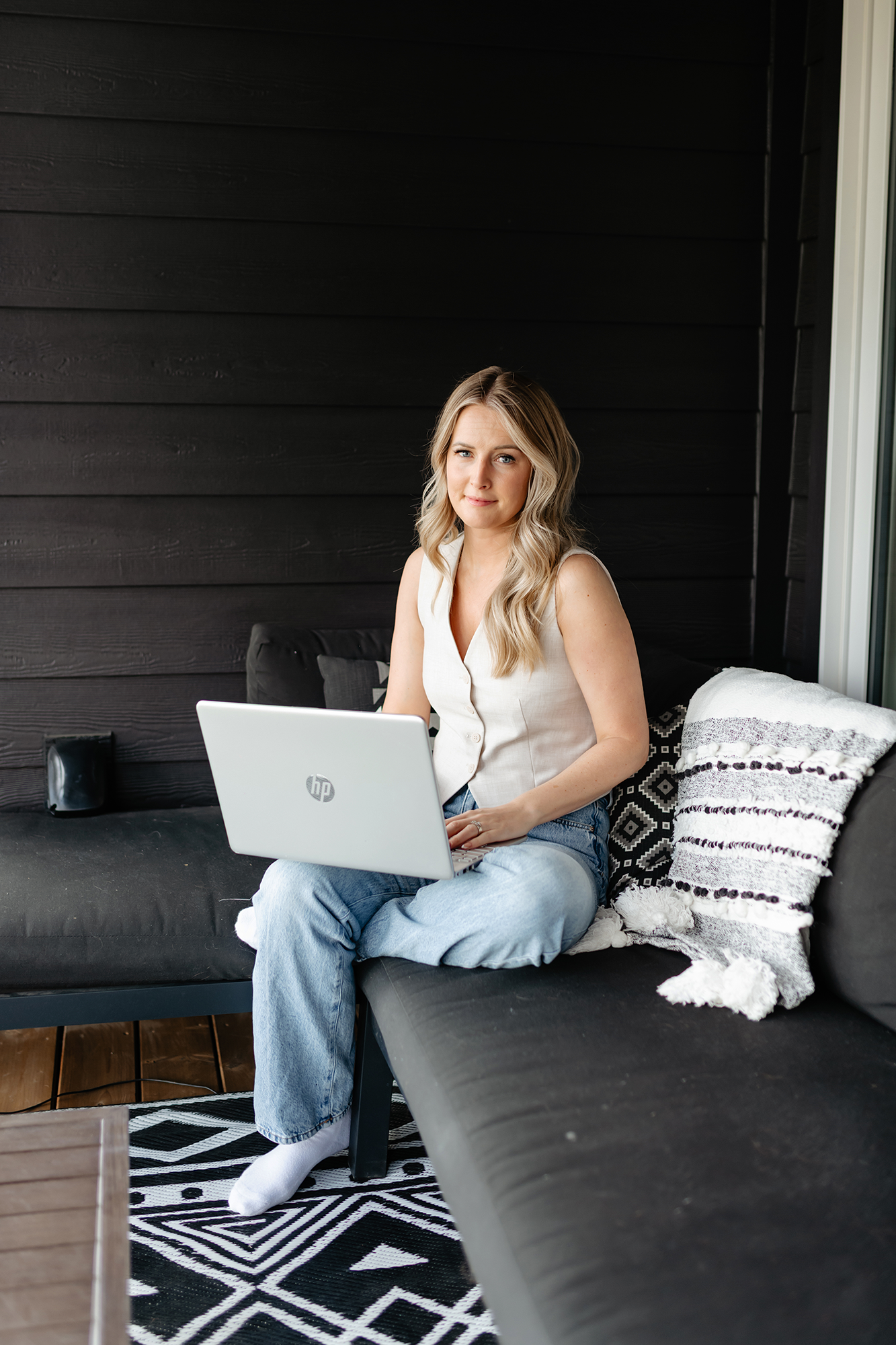 A woman with blonde hair and wearing a white sleeveless top and blue jeans, sitting on a black outdoor sofa with her laptop on her lap, in a space with black wooden walls and a geometric black and white rug. She is looking at the camera with a slight smile.