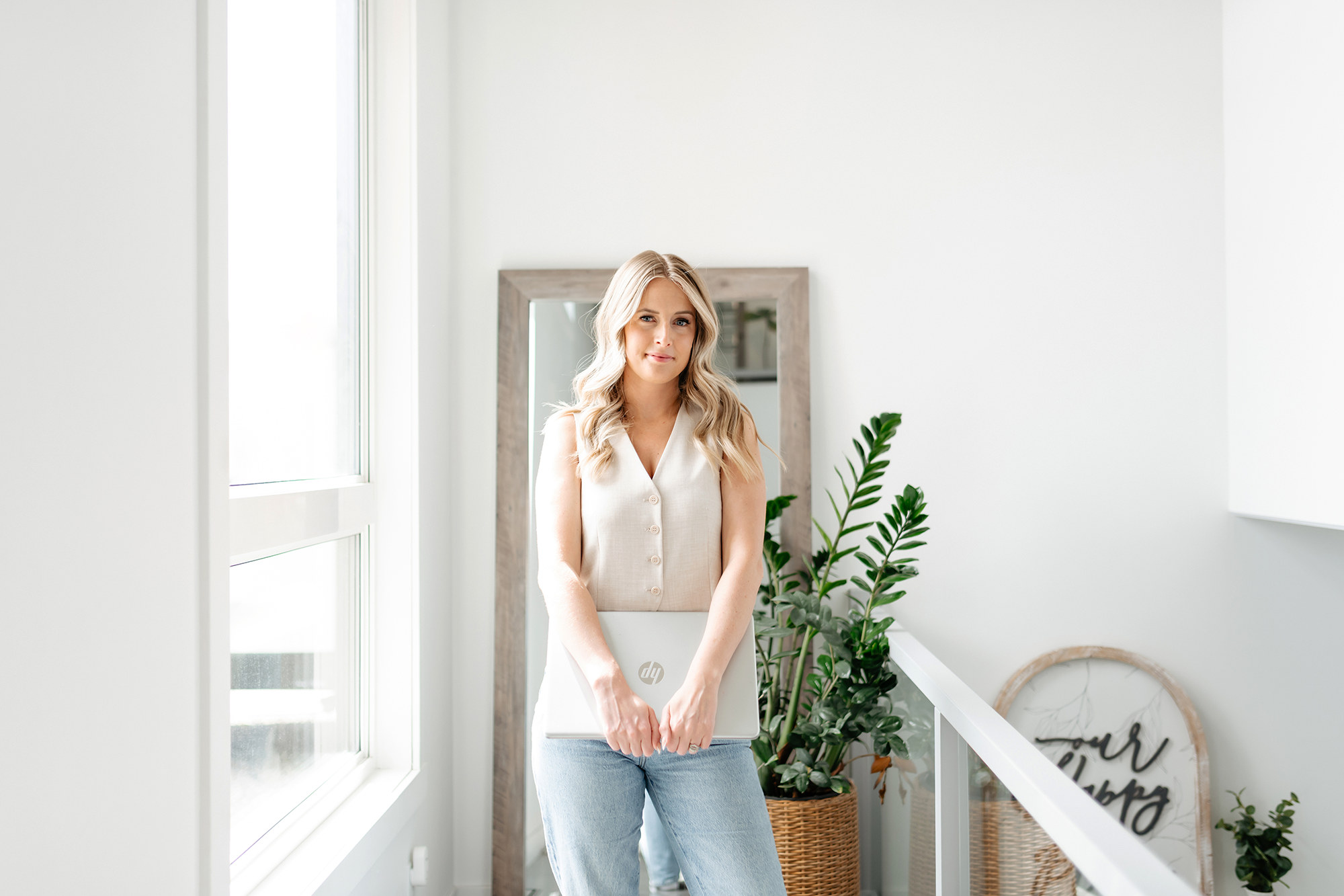 A woman with long blonde hair stands by a window, holding a closed silver HP laptop, smiling softly in a bright, modern room with a mirror, green plants, and a framed artwork in the background.