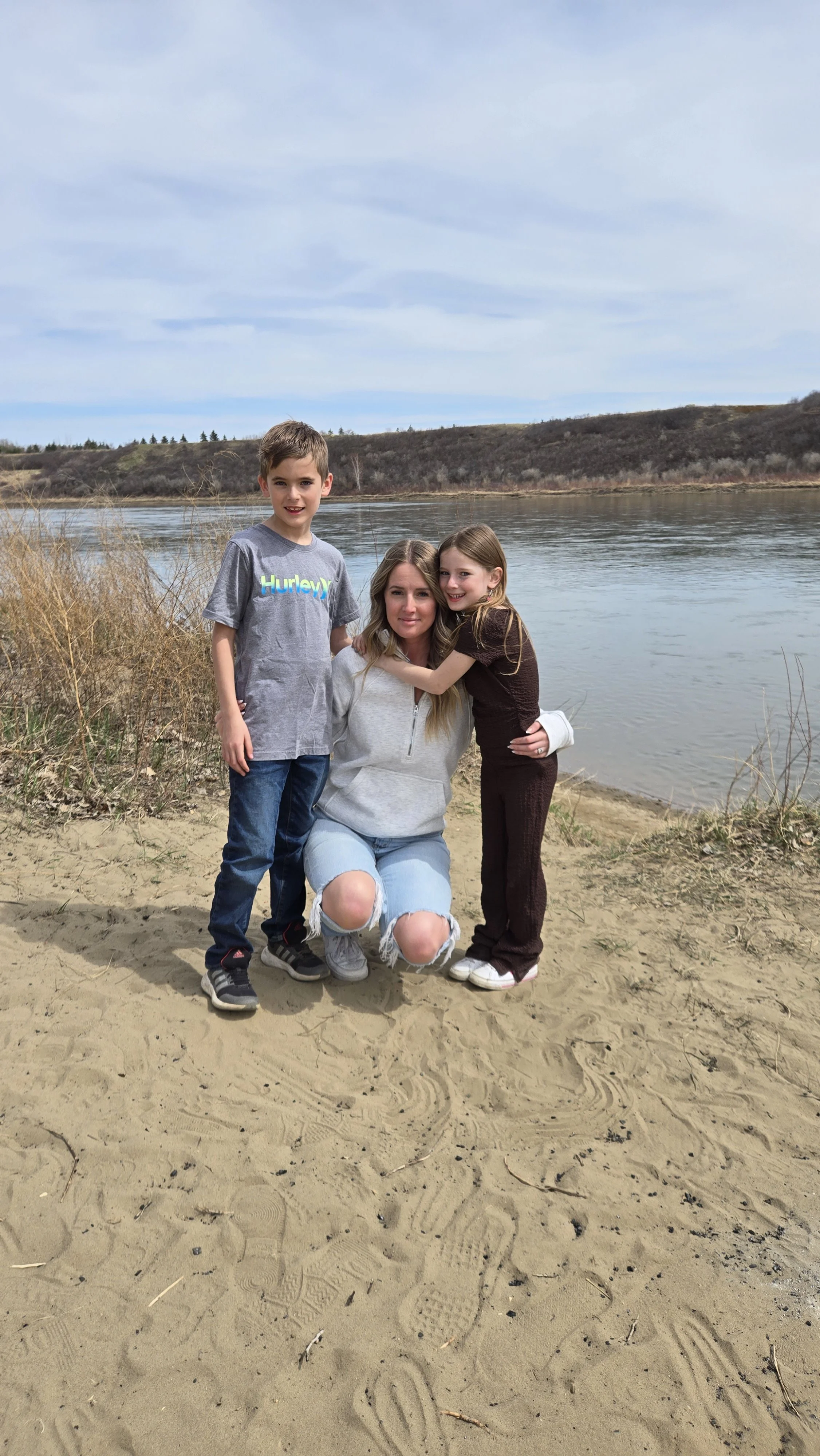 A woman kneeling on a sandy beach with two children, near a body of water and hilly landscape in the background.