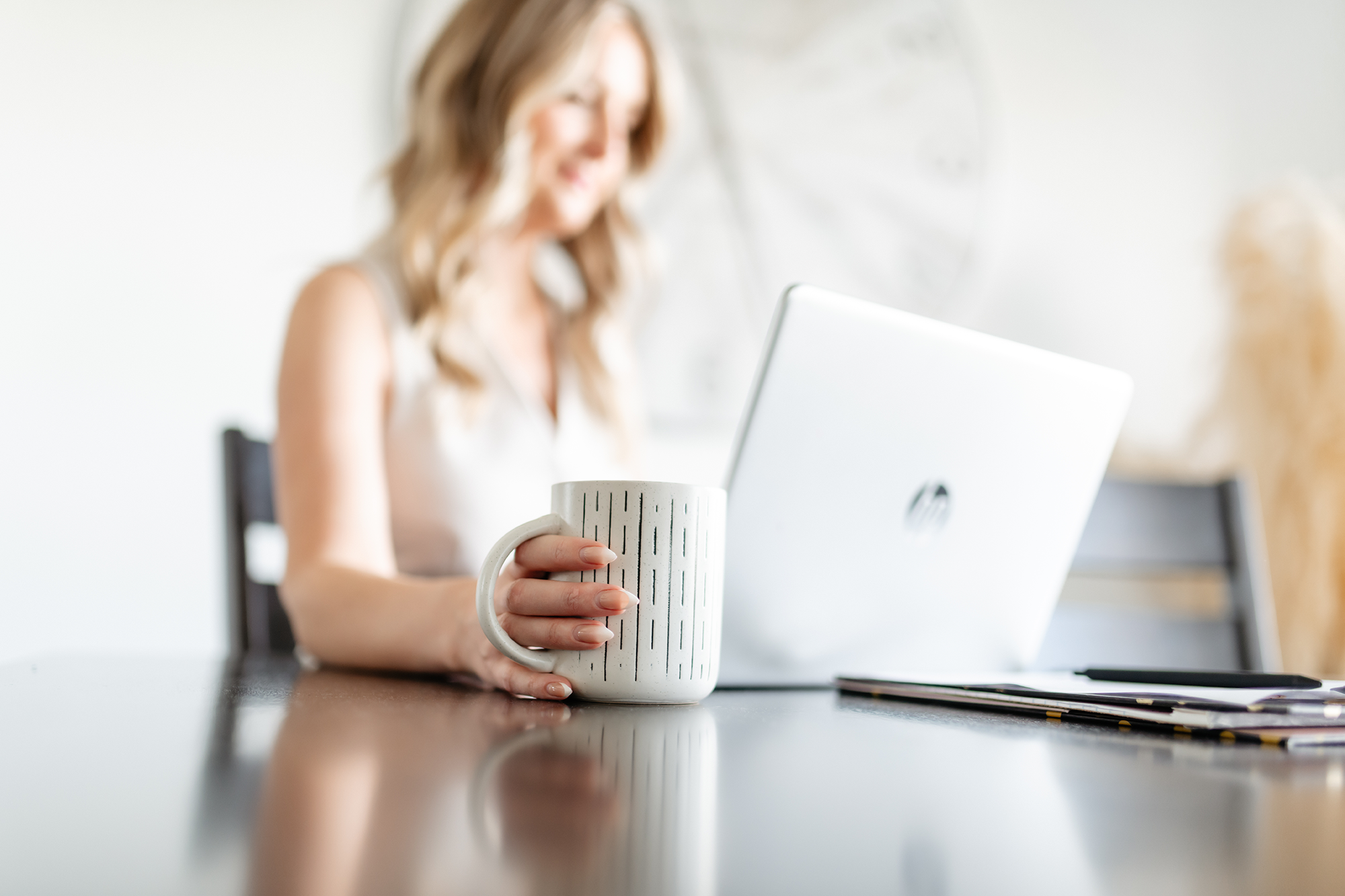 A woman with blonde hair sitting at a table, holding a white mug with vertical black stripes, in front of a silver laptop in a bright, cozy room.