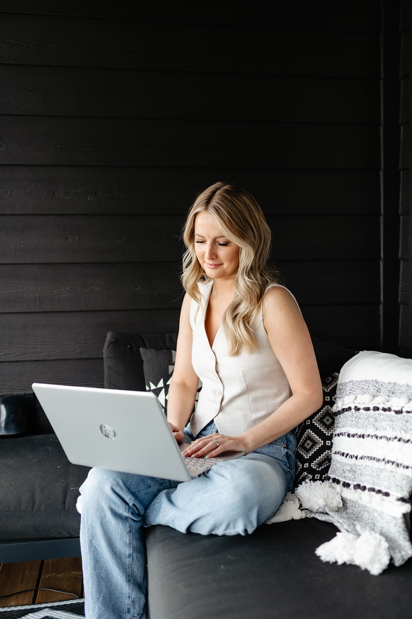 A woman with long blonde hair seated on a black sofa, working on a silver laptop, in a room with black wooden walls and decorative pillows.