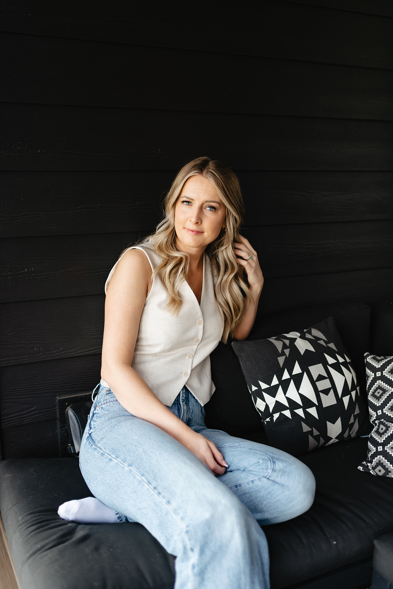 A woman with blonde wavy hair, blue eyes, wearing a white sleeveless button-up top and light blue jeans, is sitting on a black cushioned bench with decorative black and white pillows. She is lightly touching her hair and looking at the camera. The background features black wooden paneling.