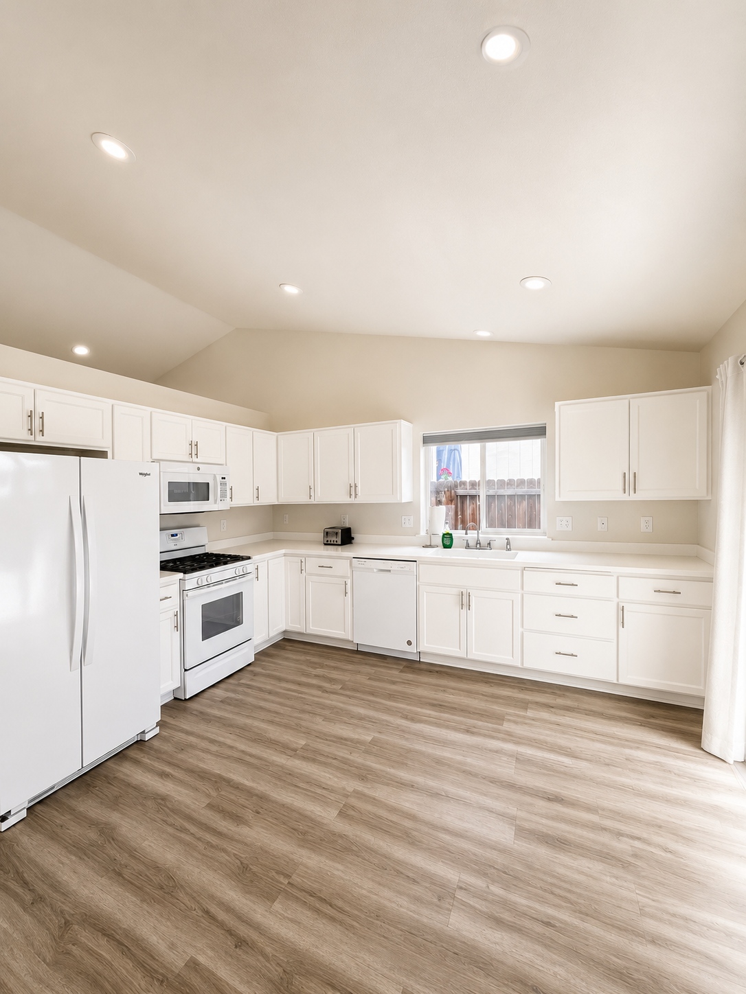 Empty kitchen with white cabinets, white appliances, a window above the sink, and wood flooring.