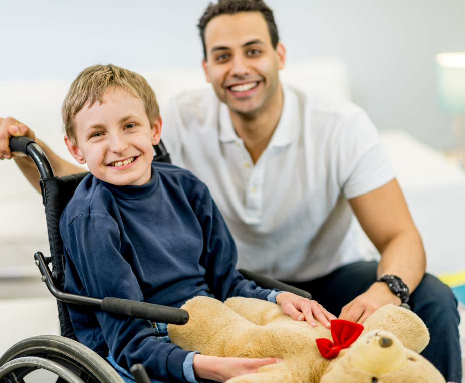 A young boy in a wheelchair holding a teddy bear with a red bow, and an adult man kneeling beside him in a bright room, both smiling.