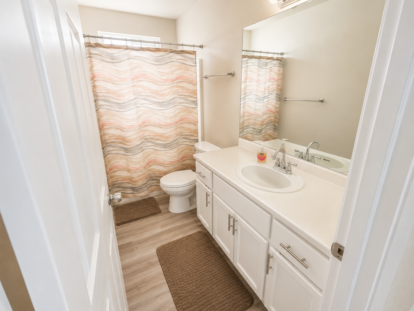 A small bathroom with a white vanity, a large mirror, a toilet, and a shower with a colorful striped curtain. There are two brown rugs on the light wood floor and a soap dispenser on the sink.