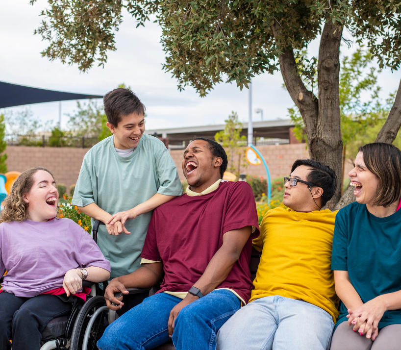 Group of five young people with disabilities outdoors under a tree, laughing and enjoying each other's company.