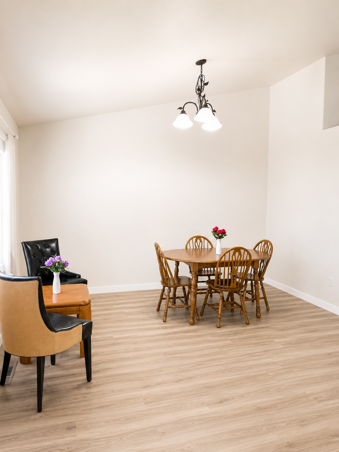 Dining room with a wooden table and four matching chairs, a black armchair and a tan armchair with a side table, all with flower vases, wood flooring, white walls, and a ceiling light fixture.