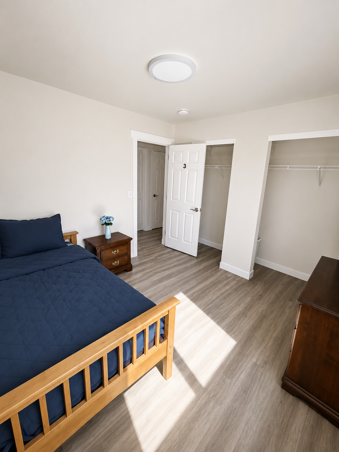 A bedroom with a wooden bed frame and navy bedding, a small nightstand with a blue vase and flowers, a closet with open doors, and a dresser.