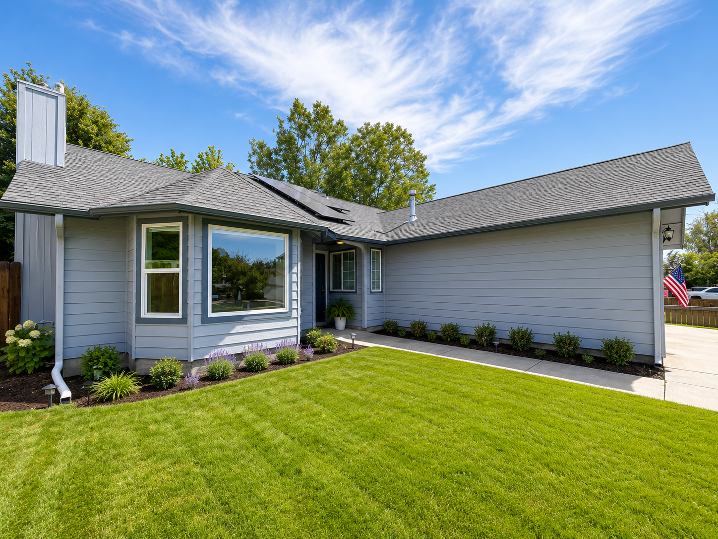 Front yard of one of our homes with green lawn, flower beds, and a light gray exterior with a large bay window.