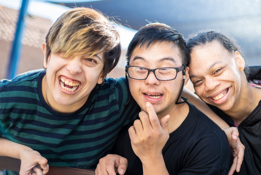 Three friends smiling and posing together outdoors, with one person wearing glasses and the others showing happy expressions.