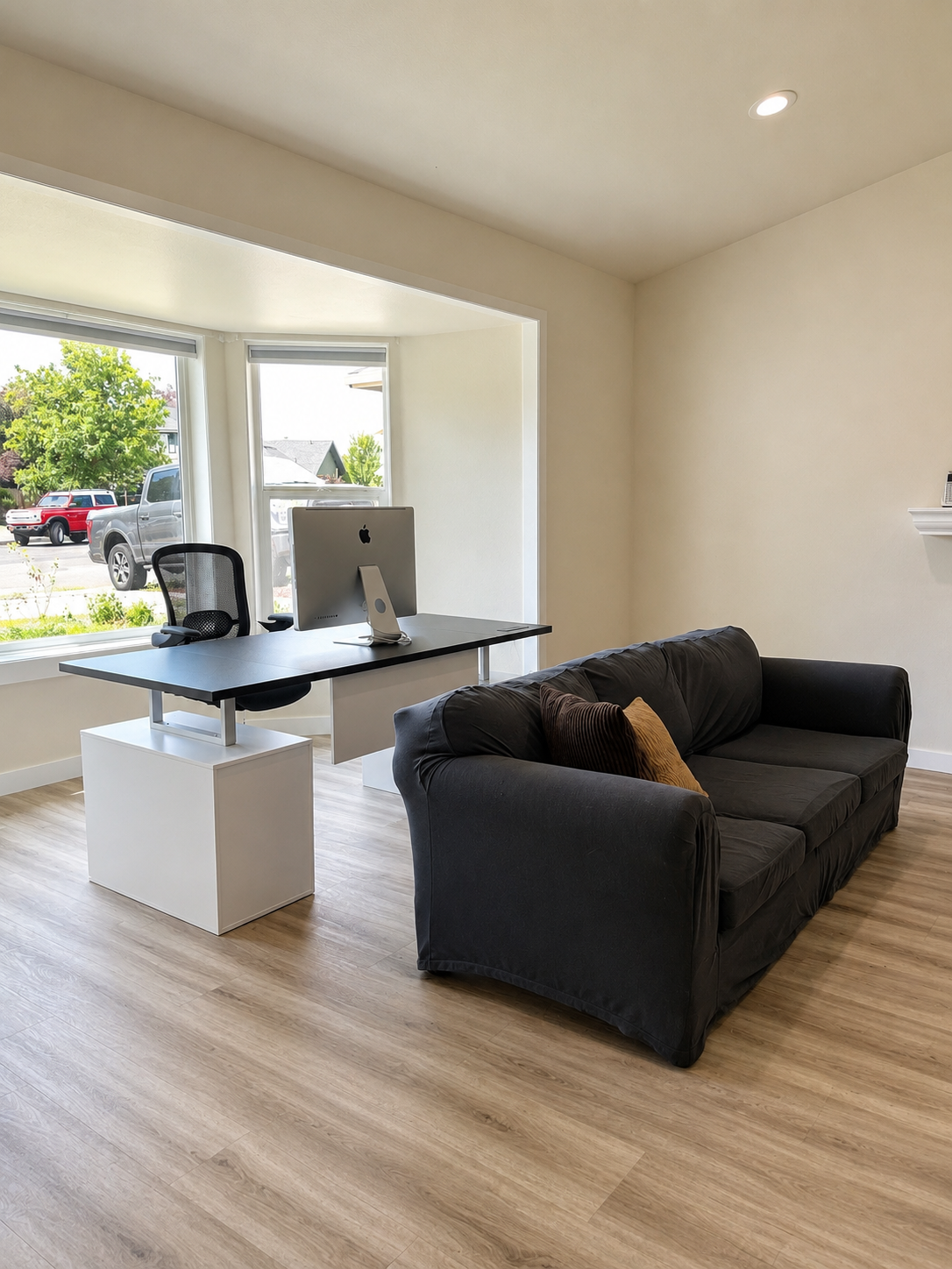 Home office with a black couch, a white desk with a computer, and large windows showing a neighborhood