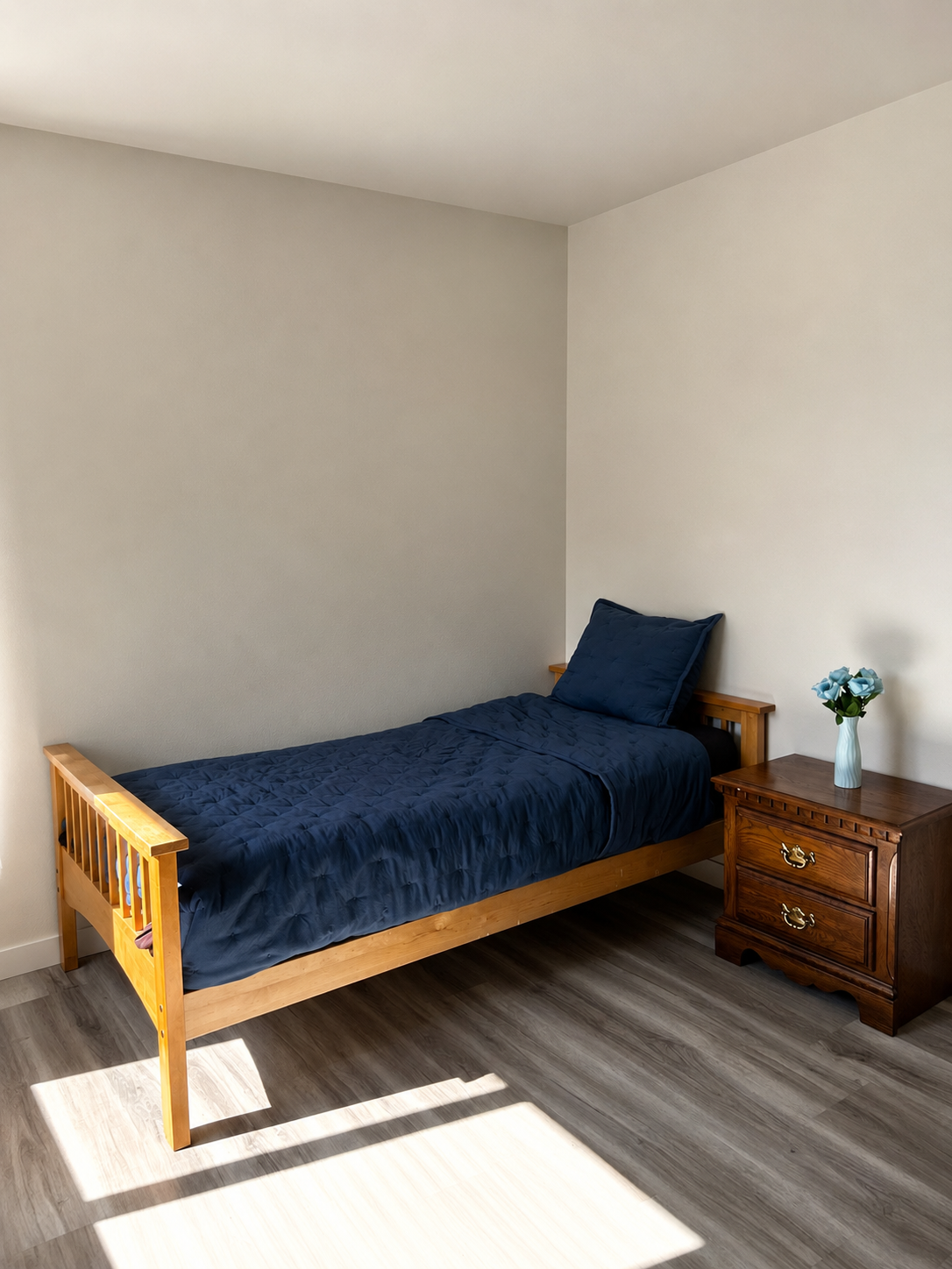 A simple bedroom with a wooden bed frame, dark blue quilt, and matching pillow. A wooden nightstand with two drawers and brass hardware holds a white vase with blue roses. The room has light-colored walls and wooden flooring, with sunlight coming in 