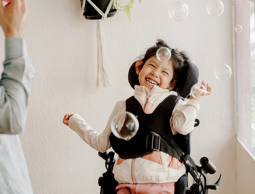 A disabled girl with dark hair in pigtails laughing happily in a stroller with bubbles floating around, indoors near a window.