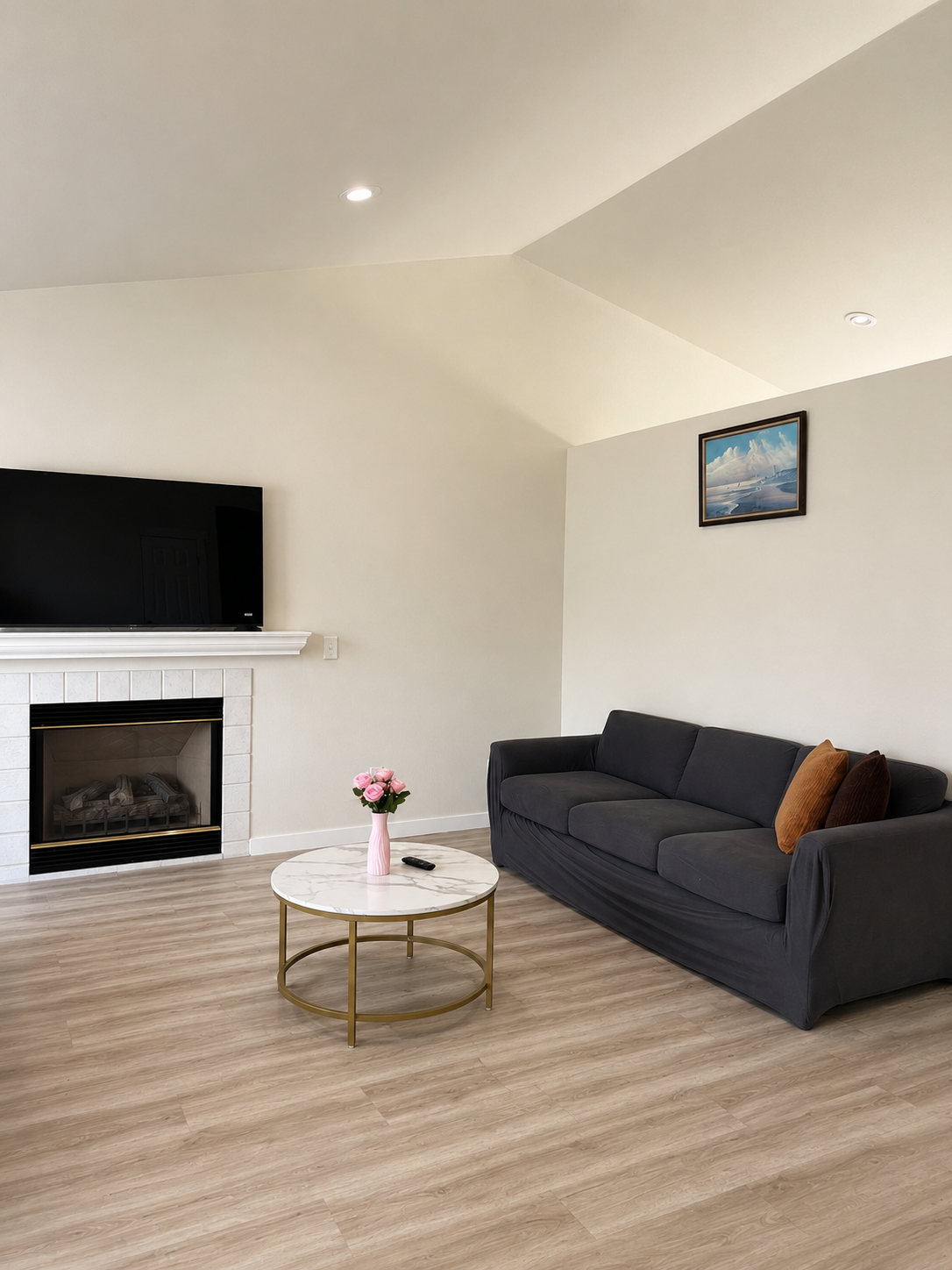 A living room with a black sofa, a small round marble coffee table with a gold frame, decorated with a pink vase of roses and a remote control. There is a white fireplace with a black television mounted above it, a small landscape painting on the wal