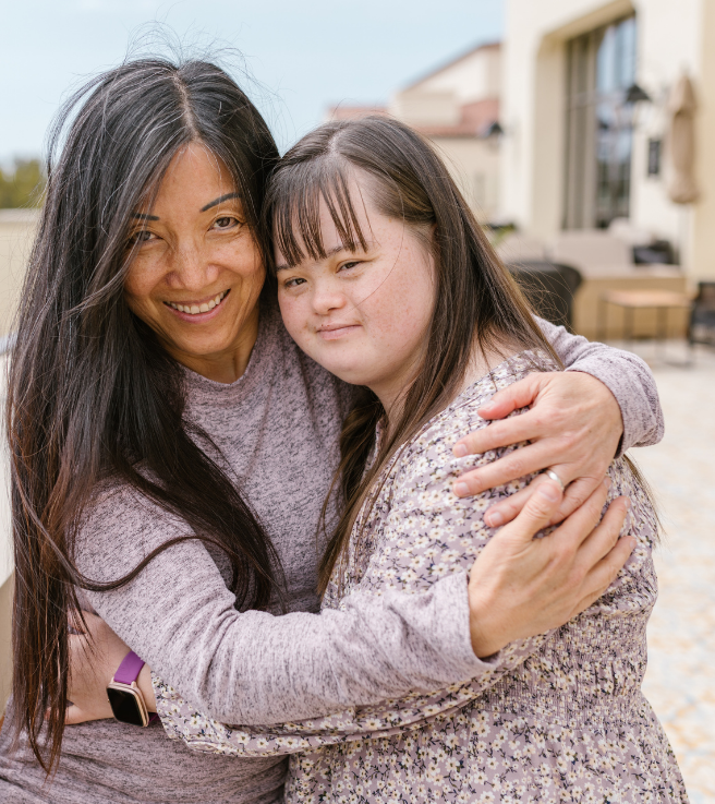 A woman and a girl hugging outdoors, smiling and looking at the camera, with a house in the background.
