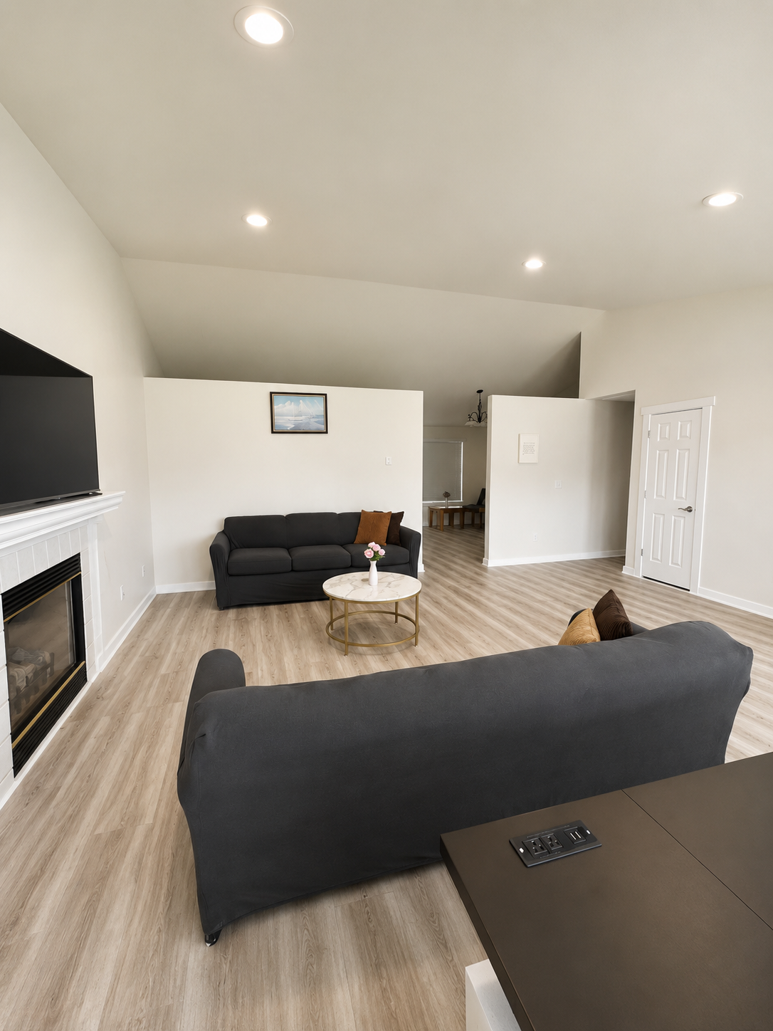 Living room with black sofas, a coffee table with pink flowers, a television on a white fireplace mantel, and a light-colored wood floor.