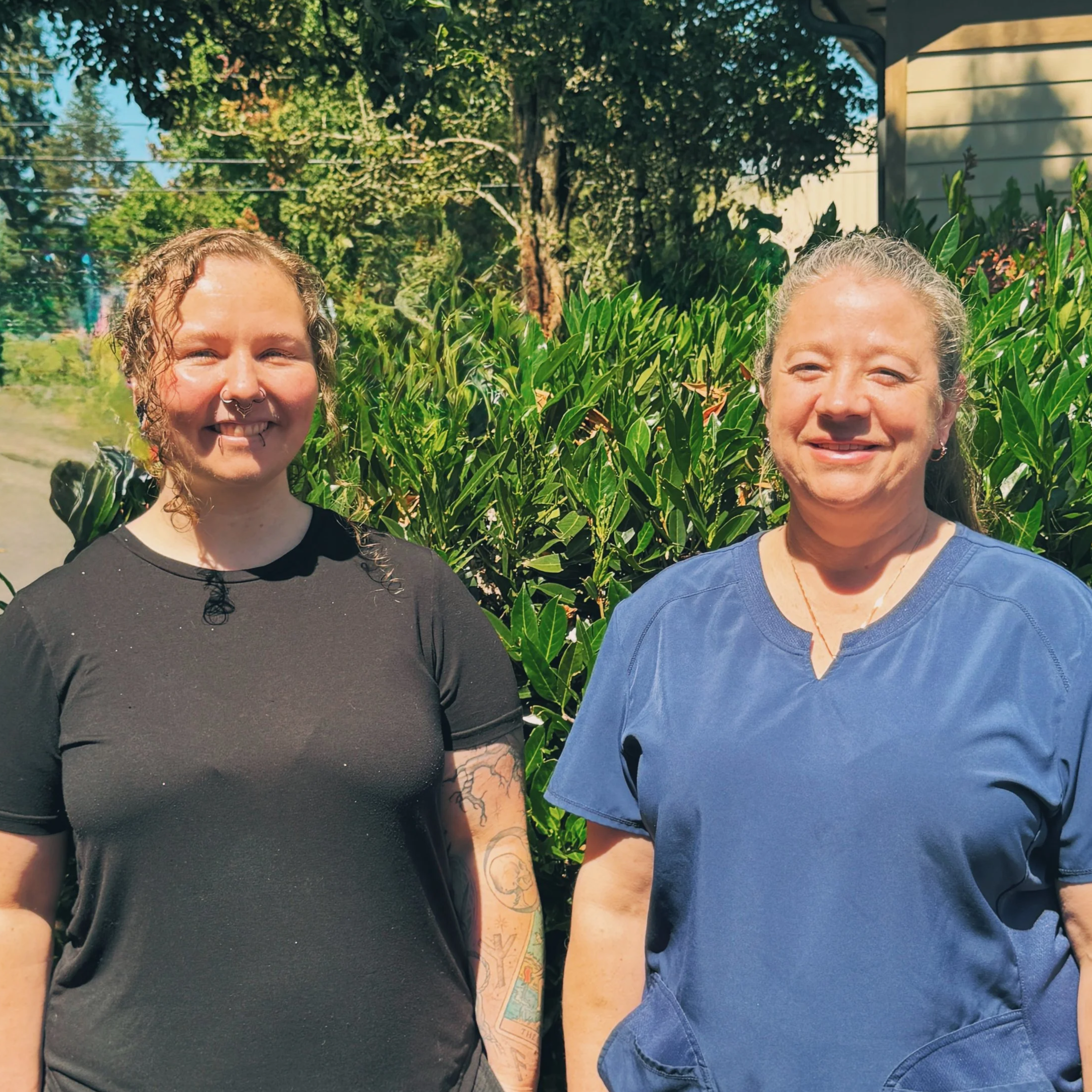 Two women smiling outdoors, surrounded by green plants and trees, wearing casual clothing.