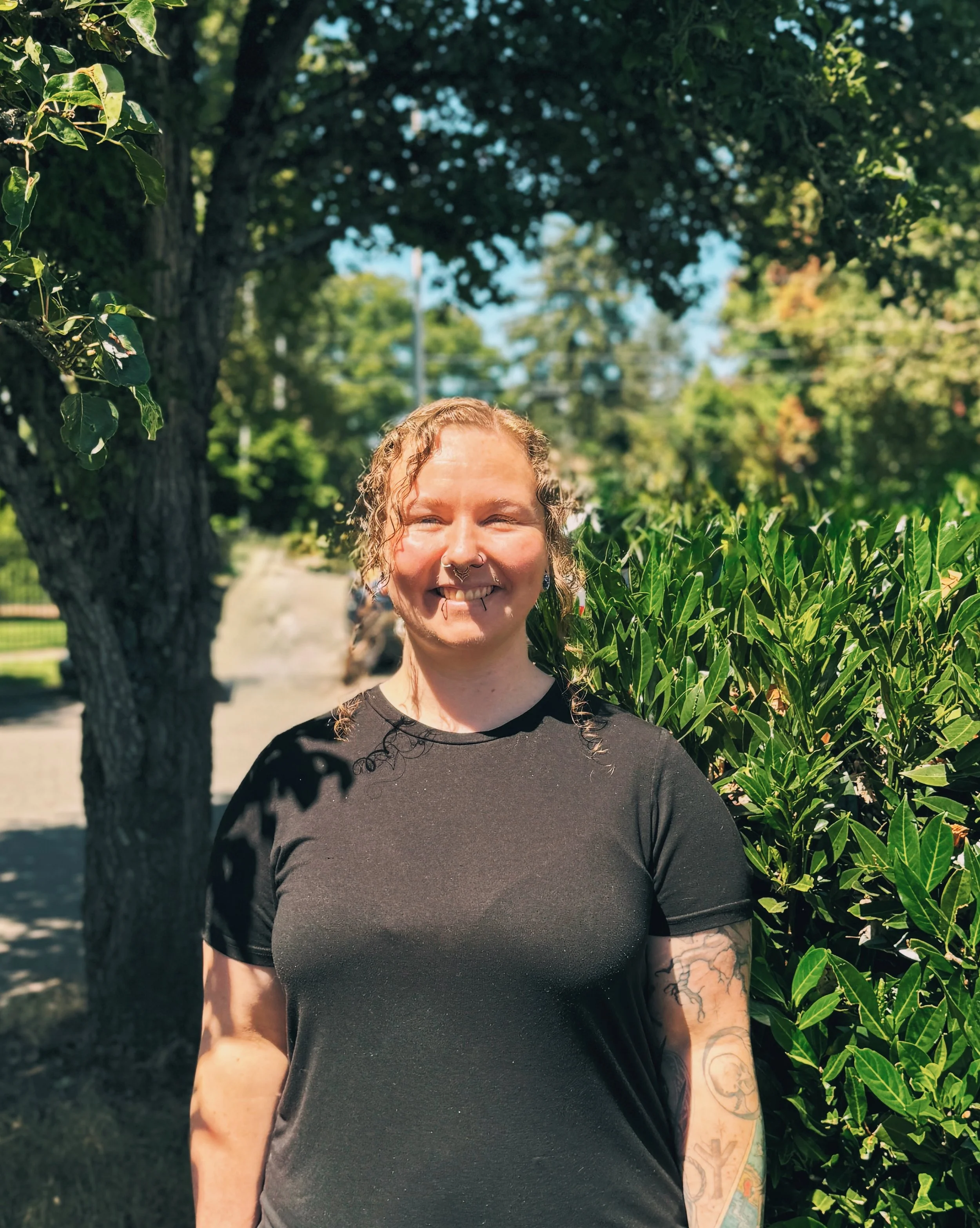 A smiling woman with tattoos on her arm, wearing a black t-shirt, standing outdoors near green bushes and trees on a sunny day.