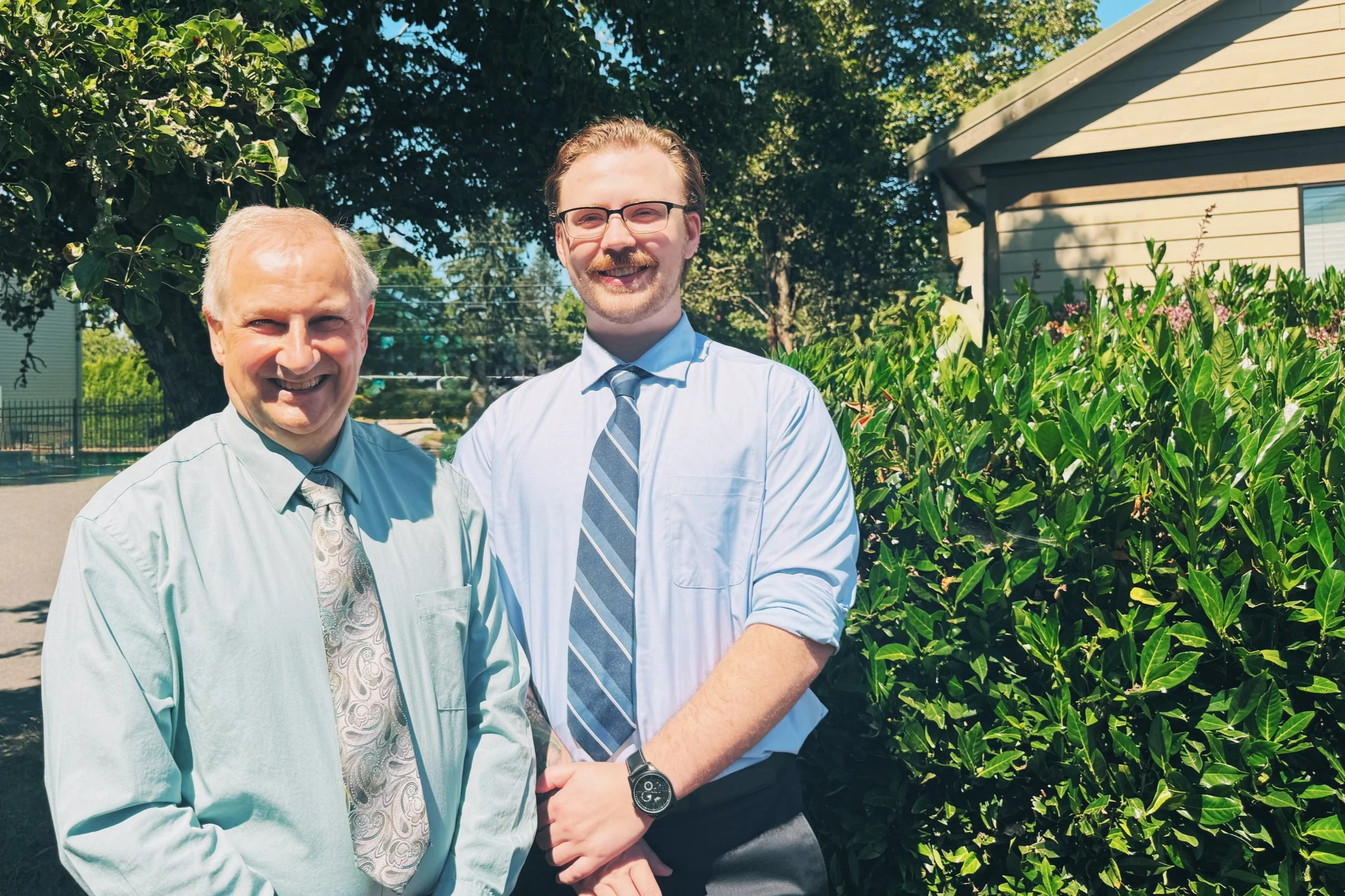 Two men wearing dress shirts and ties, smiling outdoors, with greenery and a house in the background.