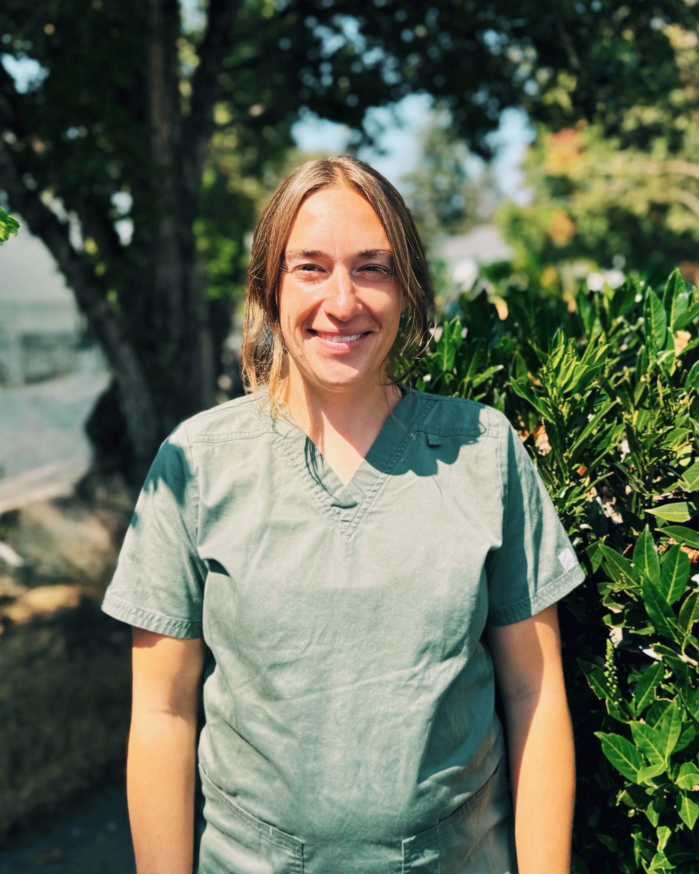 A woman with long brown hair smiling outdoors near green foliage and trees, wearing a light green hospital or medical scrubs top.