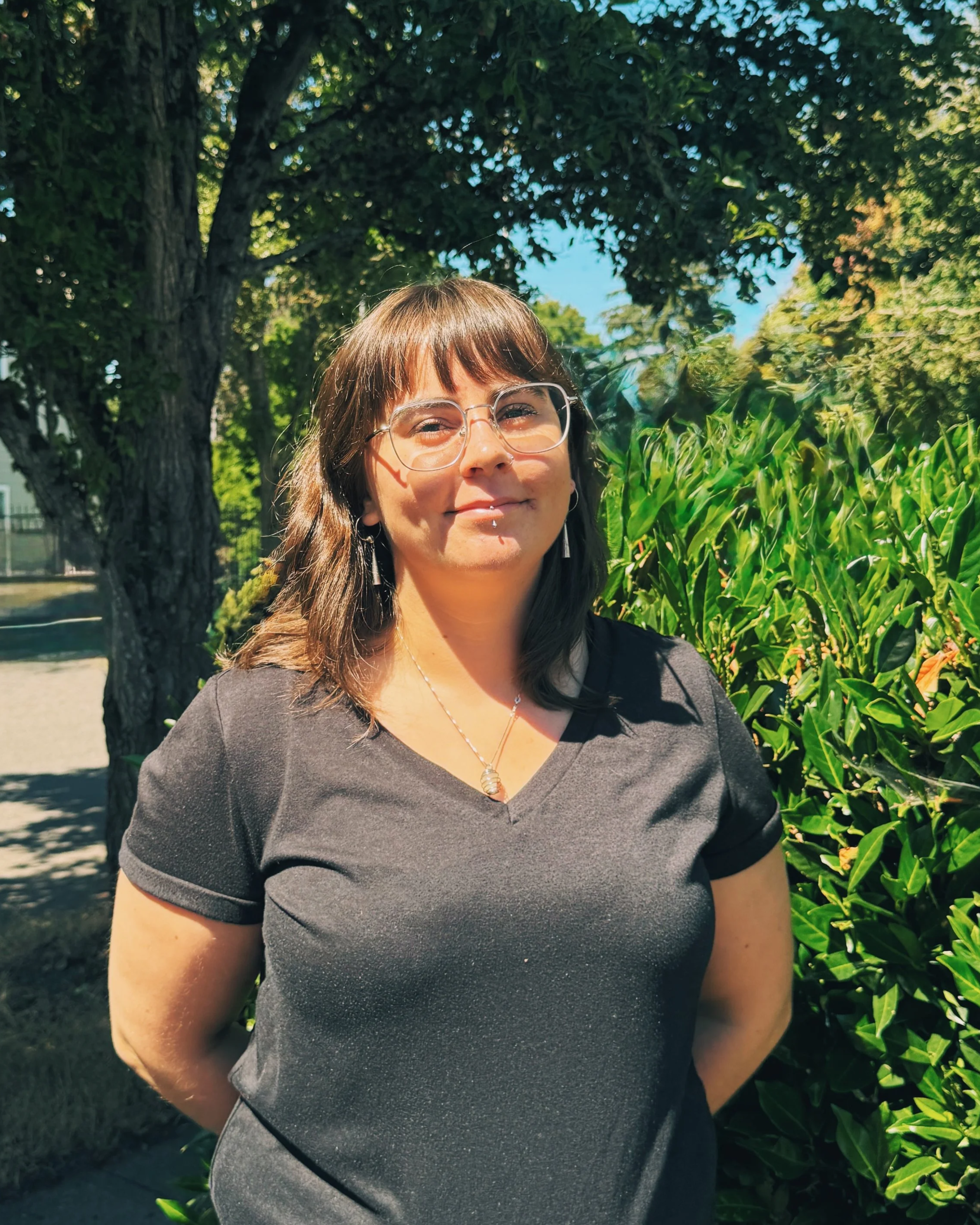 A woman with brown hair and glasses standing outdoors near green plants and a large tree in sunlight.