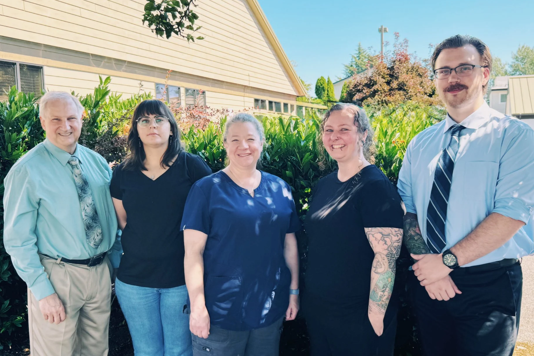 Group of five diverse people standing outdoors in front of green bushes and trees, with houses and clear blue sky in background.