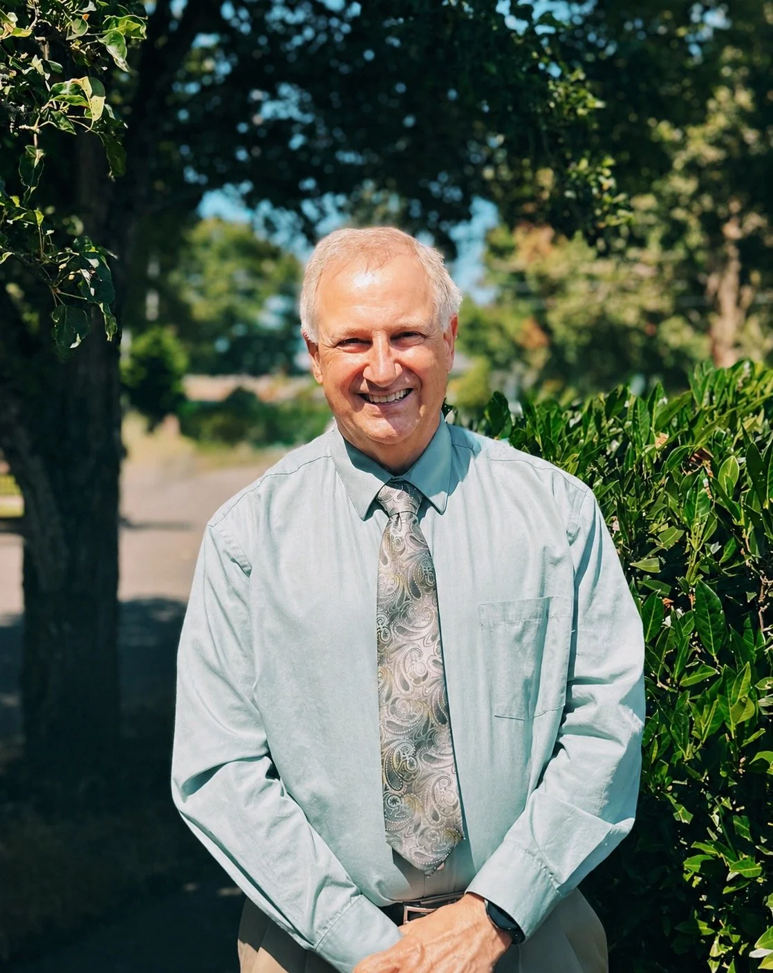 A smiling older man with light hair, wearing a light blue dress shirt and a patterned tie, standing outdoors in front of green bushes and trees, sunlight on his face.