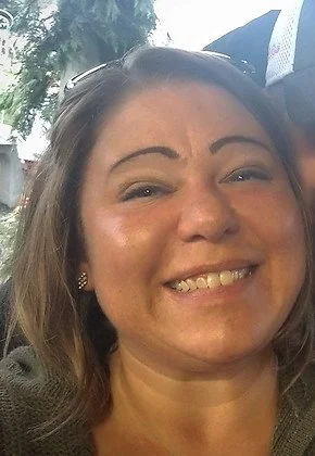 Close-up of a smiling woman with brown hair, wearing earrings, outdoors with trees and sky in the background.