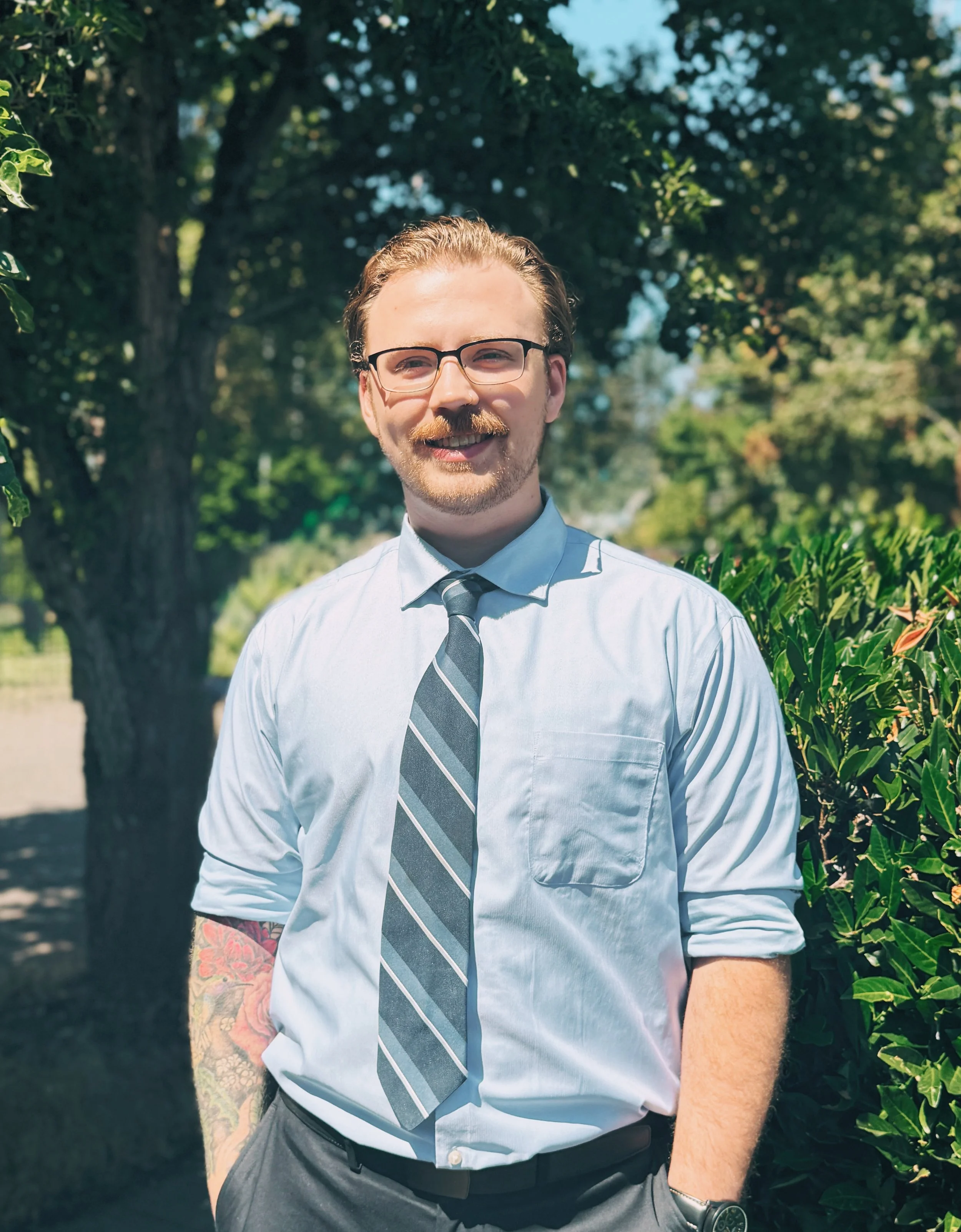 A man with glasses, a beard, and a mustache is smiling outdoors in front of trees and bushes, wearing a light blue dress shirt, a striped tie, and gray pants.