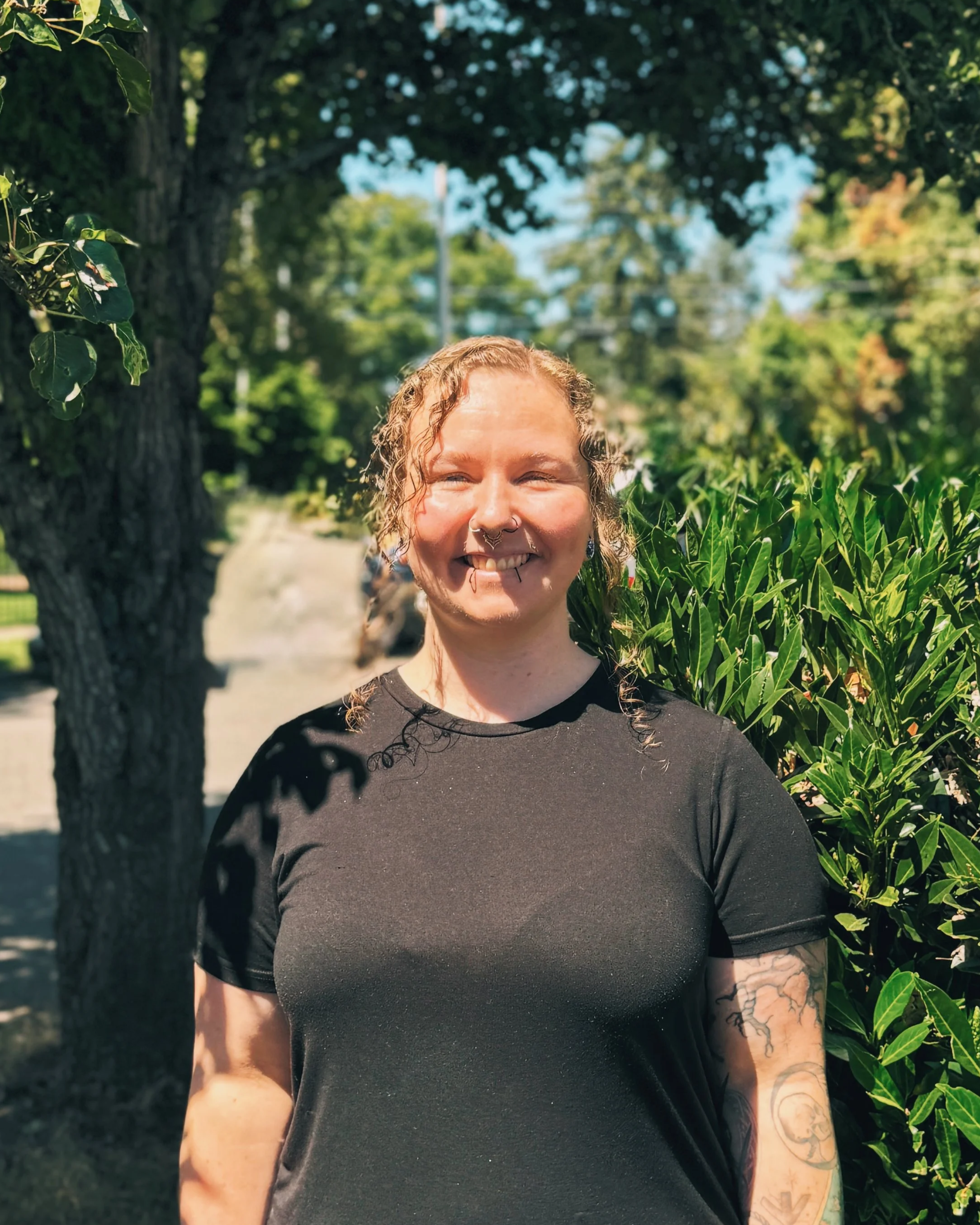 Close-up of a smiling woman with curly hair, tattoos on her arm, and a septum piercing, standing outdoors next to green plants and trees on a sunny day.