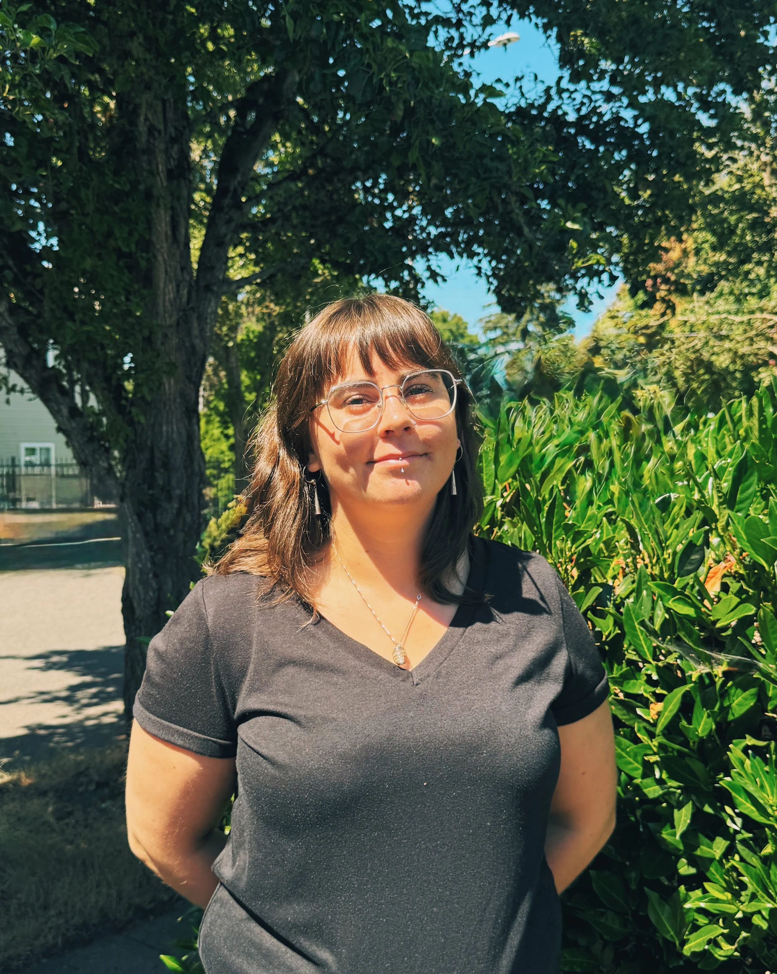A woman with brown hair and glasses standing outdoors in front of a large tree and green bushes, wearing a dark gray top, smiling slightly.
