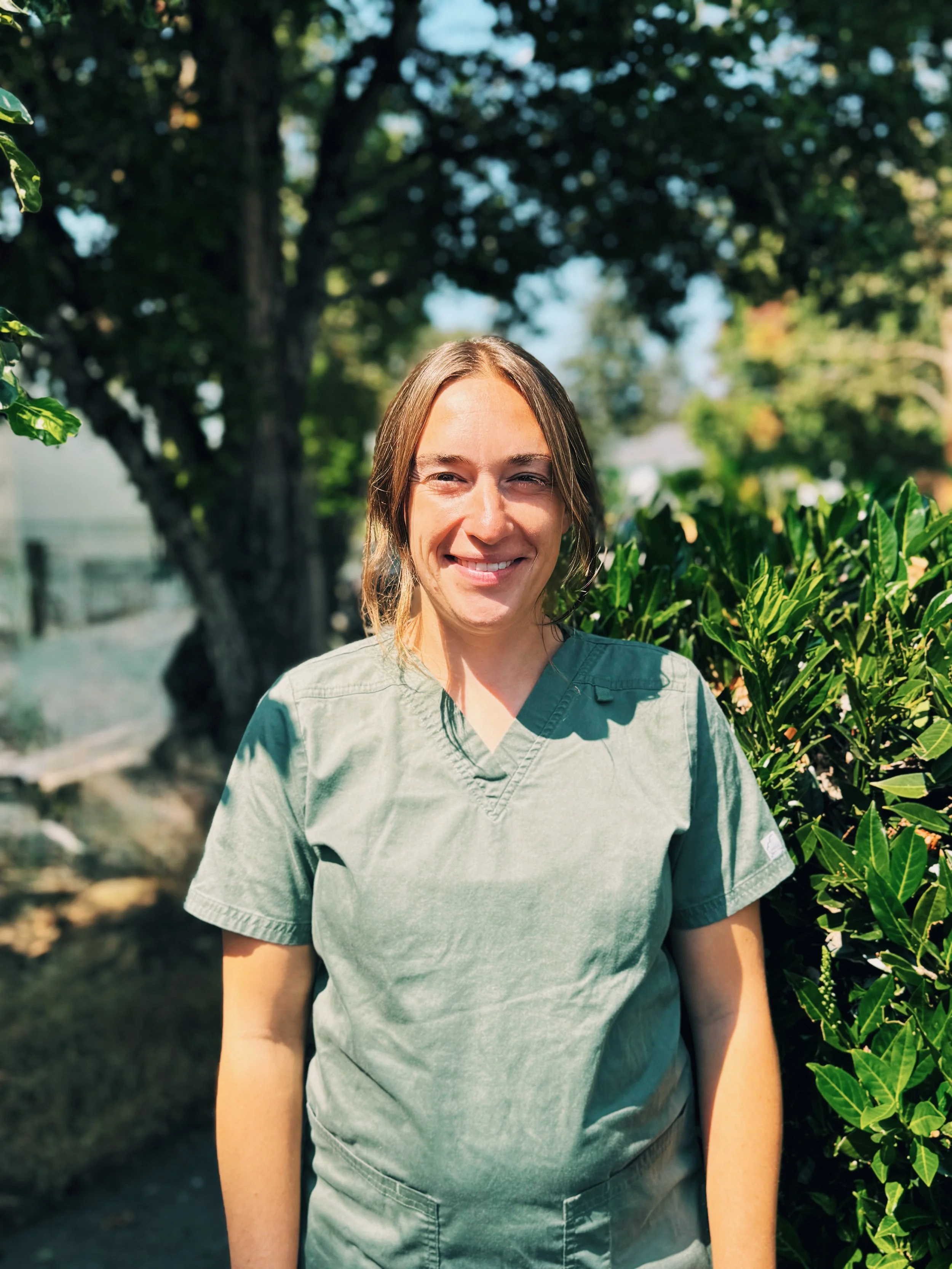 A woman with light brown hair smiling outdoors in a green uniform, standing next to leafy bushes with a large tree in the background.