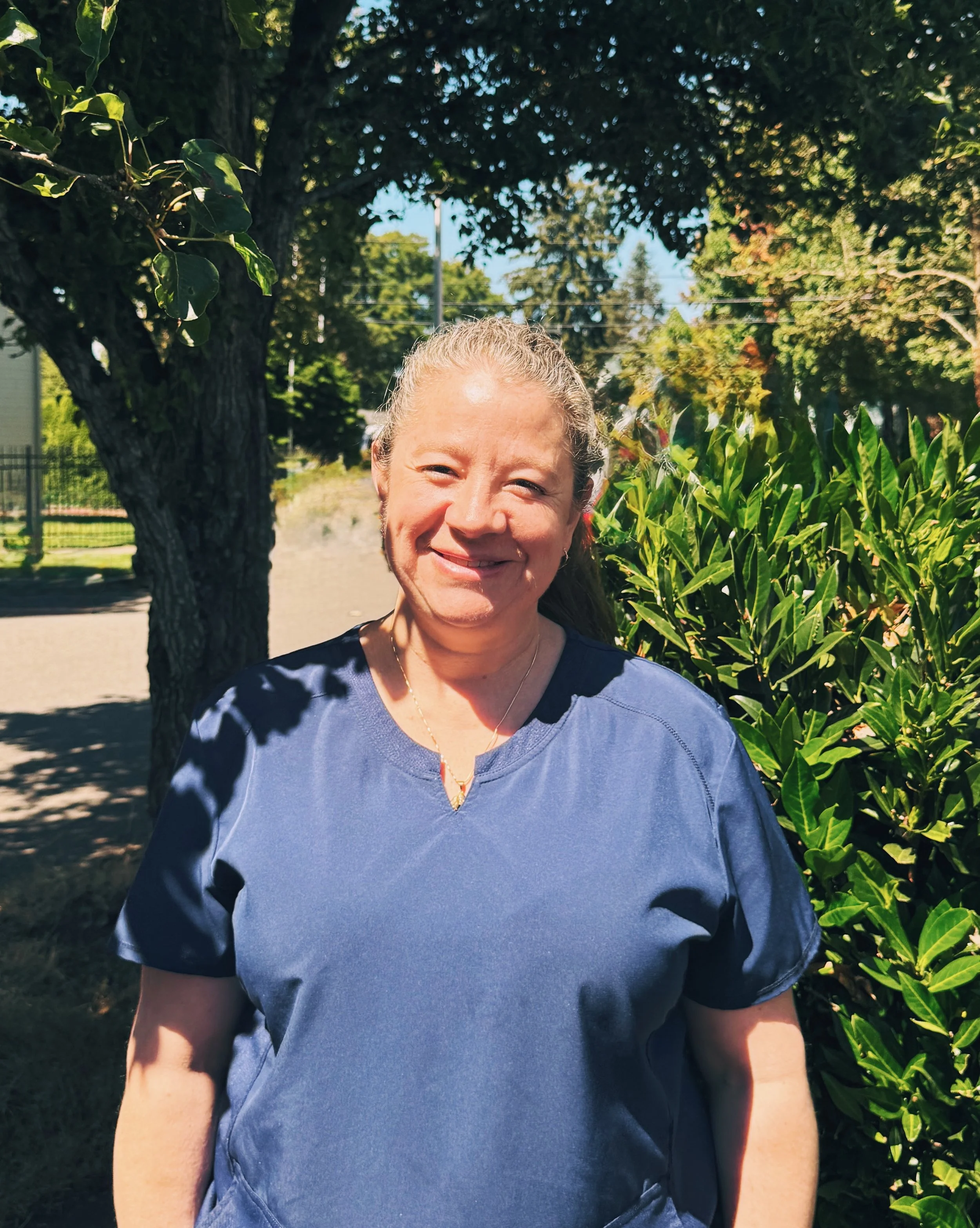 A woman smiling outdoors in a blue shirt, with greenery and trees in the background on a sunny day.