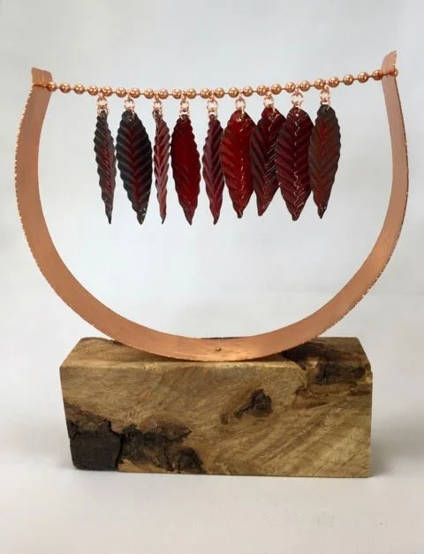 A decorative piece featuring dark red and black feathers hanging from a string of small beads, mounted on a half circle stand supported by a wooden base.
