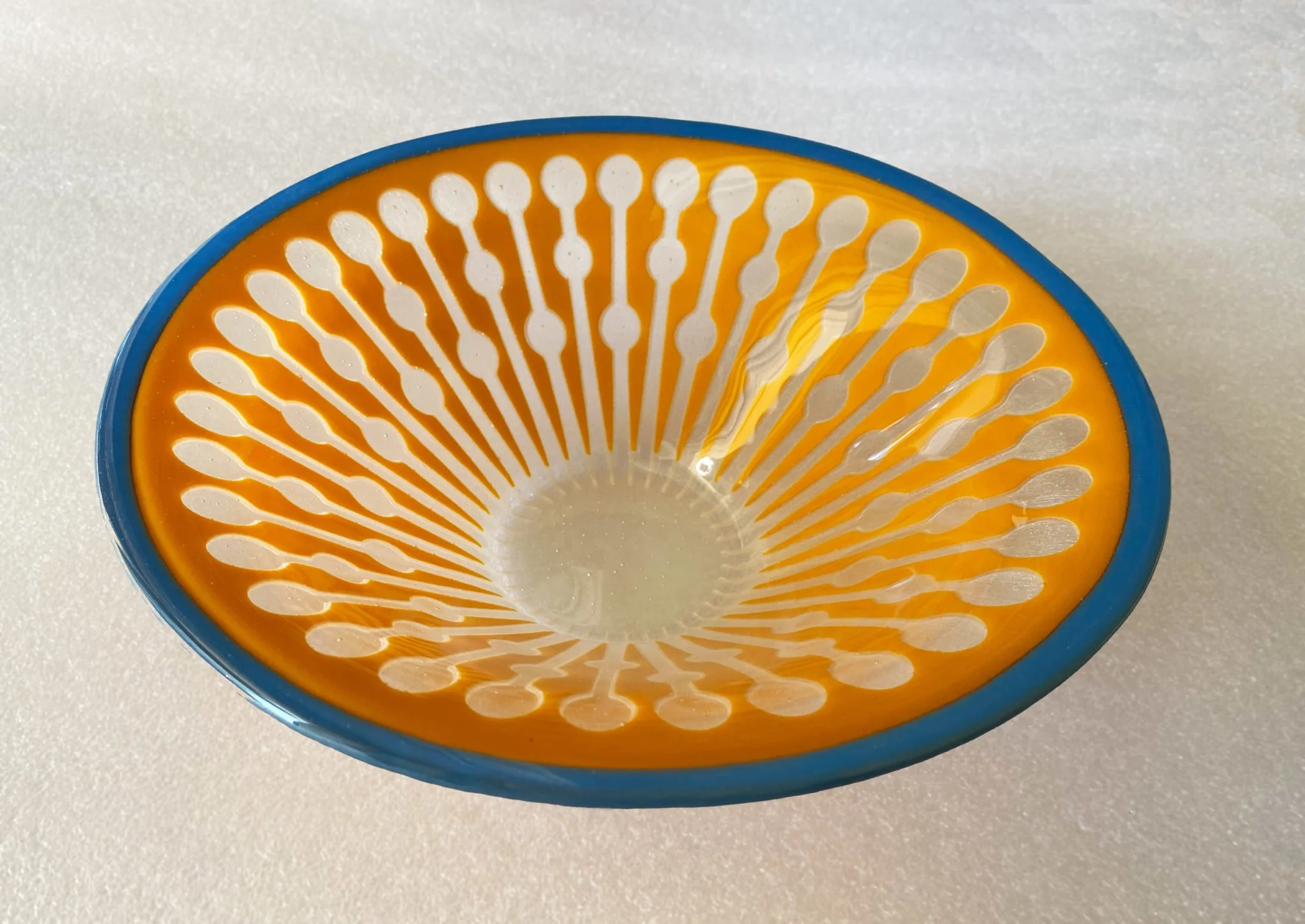 Close-up of a decorative ceramic bowl with an orange interior pattern of radiating white lines and a blue rim, placed on a light-colored surface.