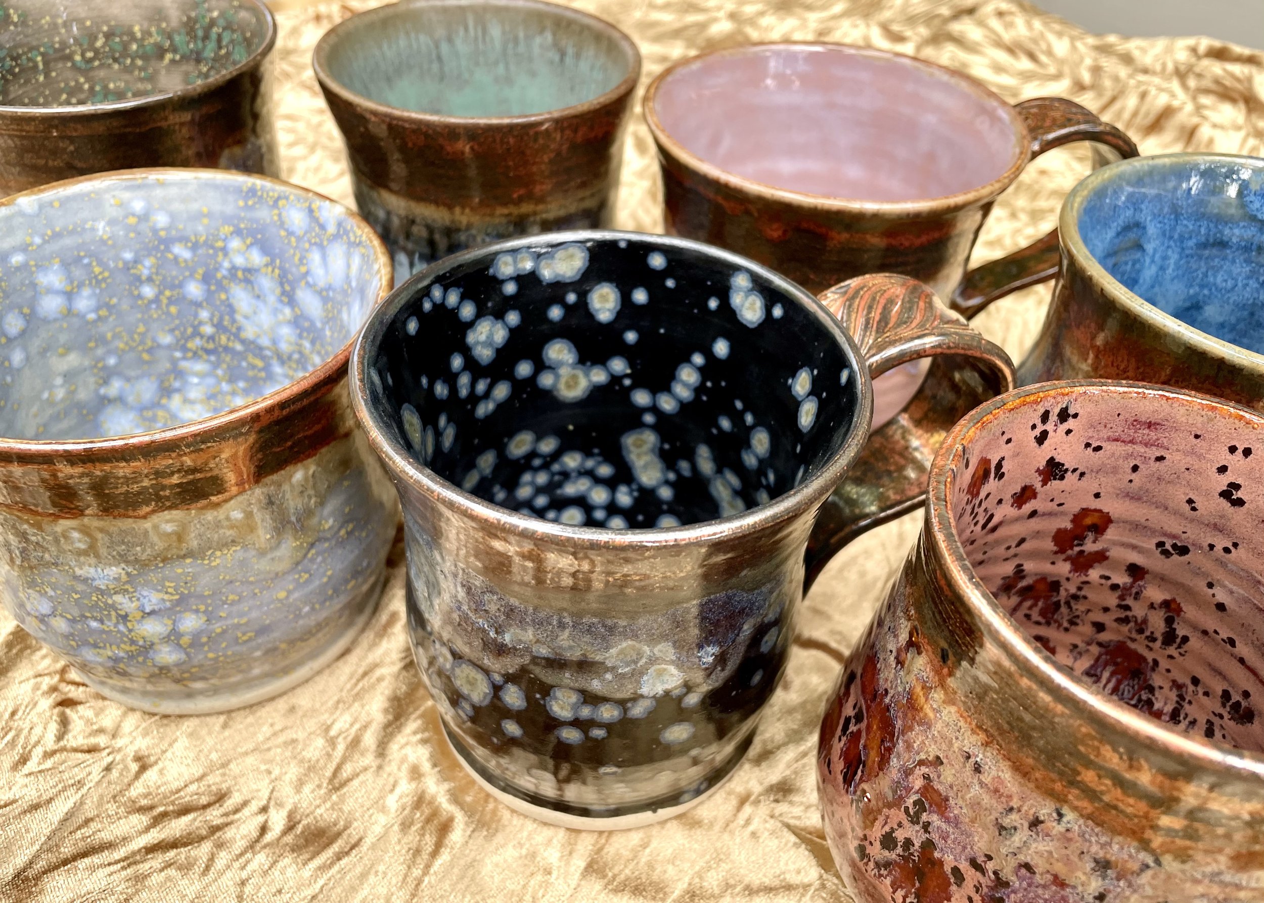 A collection of hand-crafted ceramic mugs with various glazed colors and patterns, arranged on a textured fabric background.