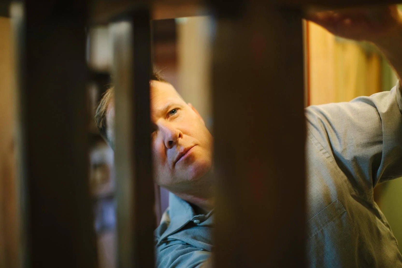 A man with light skin and short blonde hair looking through a narrow gap in a wooden bookshelf.