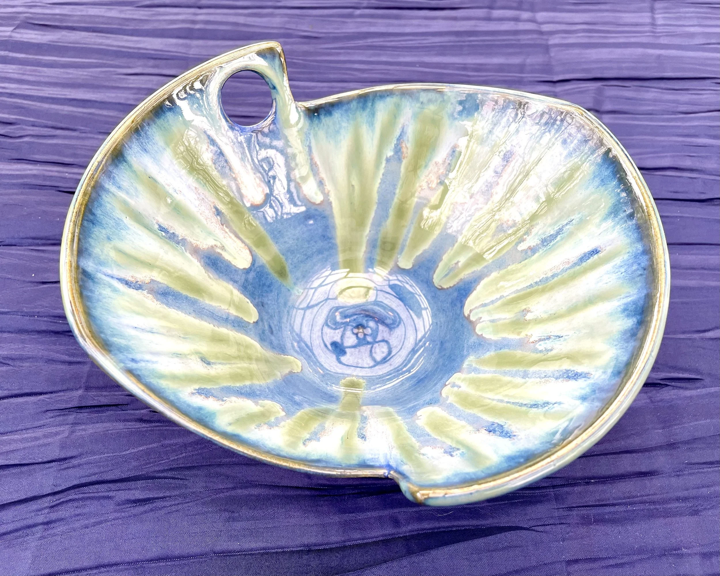A decorated ceramic spoon rest with a floral pattern in shades of blue, green, and yellow, sitting on a purple textured surface.