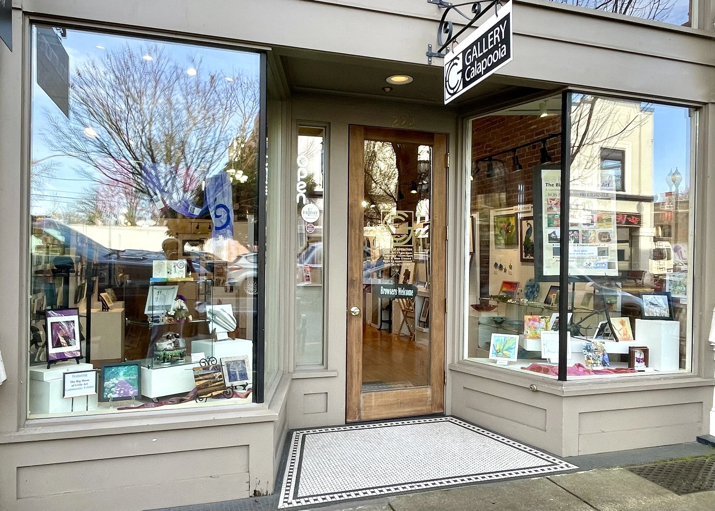 Front of an art gallery storefront with large glass windows displaying paintings and artwork inside. There is a sign above that reads 'GALLERY Calapooia' and a door with a small sign next to it. The window reflections show trees and buildings across the street.