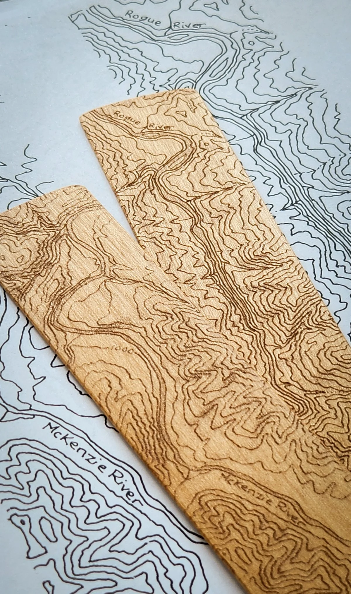 Wooden map tool with engraved contour lines of a river, placed over a printed topographic map.