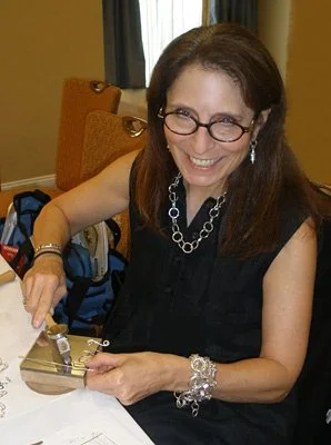 A woman with long brown hair, glasses, and jewelry, smiling as she cuts a cake in a decorated room.