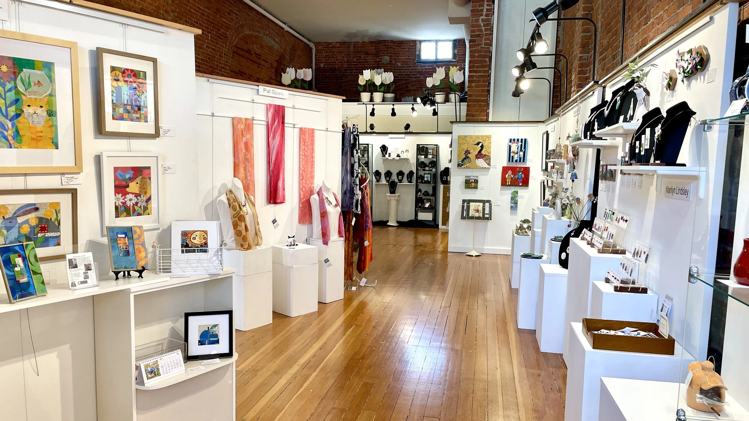 An art gallery with framed artwork on white walls, display tables of jewelry, scarves, and small art pieces, and a brick and wood interior with natural and track lighting.