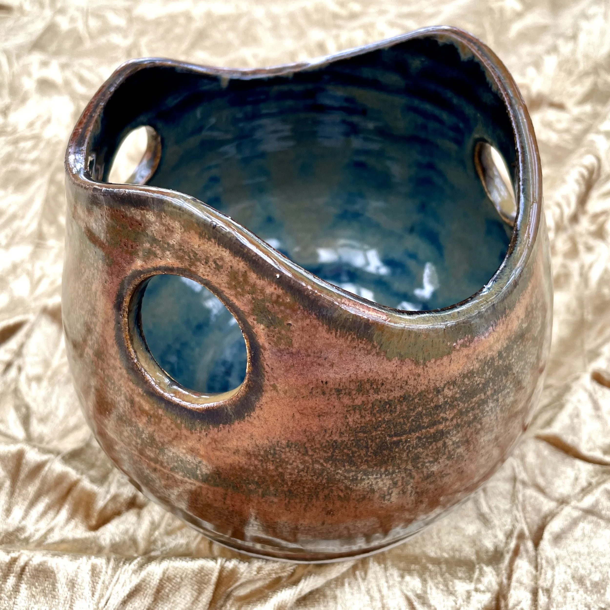 Close-up of a ceramic mug with a rustic, glossy finish, showing a wavy rim and a partially visible handle, placed on a textured beige fabric surface.
