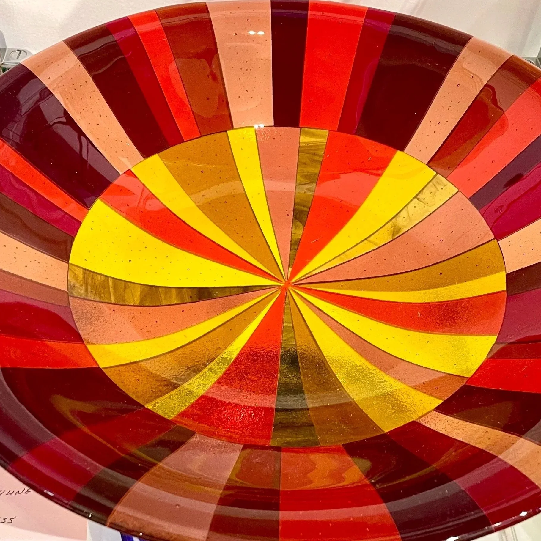 Colorful glass art piece with concentric circles and radiating segments in shades of yellow, orange, red, brown, and dark purple.