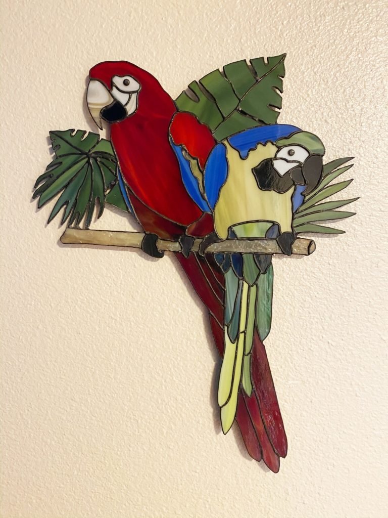 Colorful stained glass art of two parrots perched on a branch with green leaves, mounted on a beige wall.